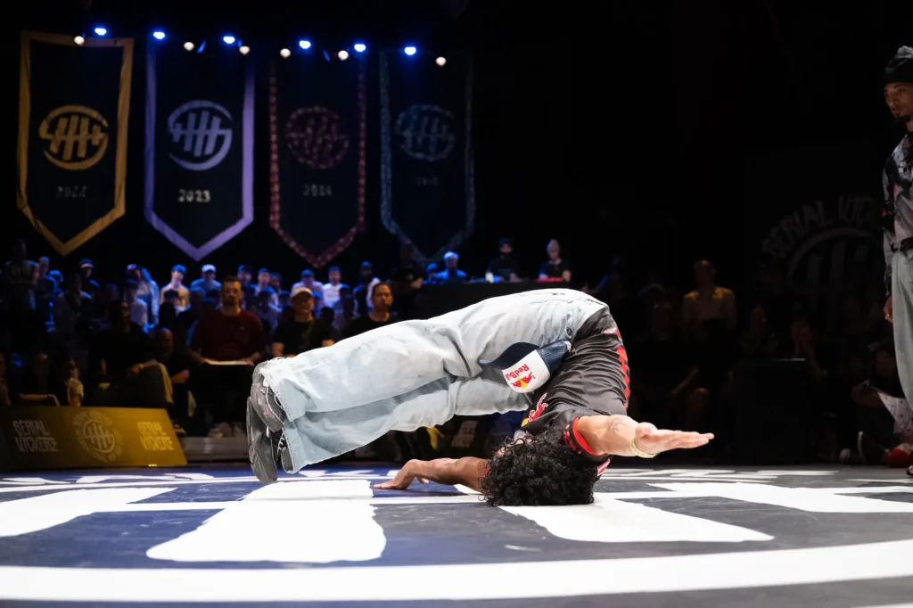 Un danseur de breakdance exécute un mouvement acrobatique où il équilibre son corps en arc sur une main, la tête et l’avant-bras posés au sol. Il porte un t-shirt noir avec des motifs rouges, un jean clair et des baskets noires. Sur sa hanche est attaché un petit sac Red Bull. En arrière-plan, un public nombreux observe attentivement la performance dans une salle sombre décorée de bannières.