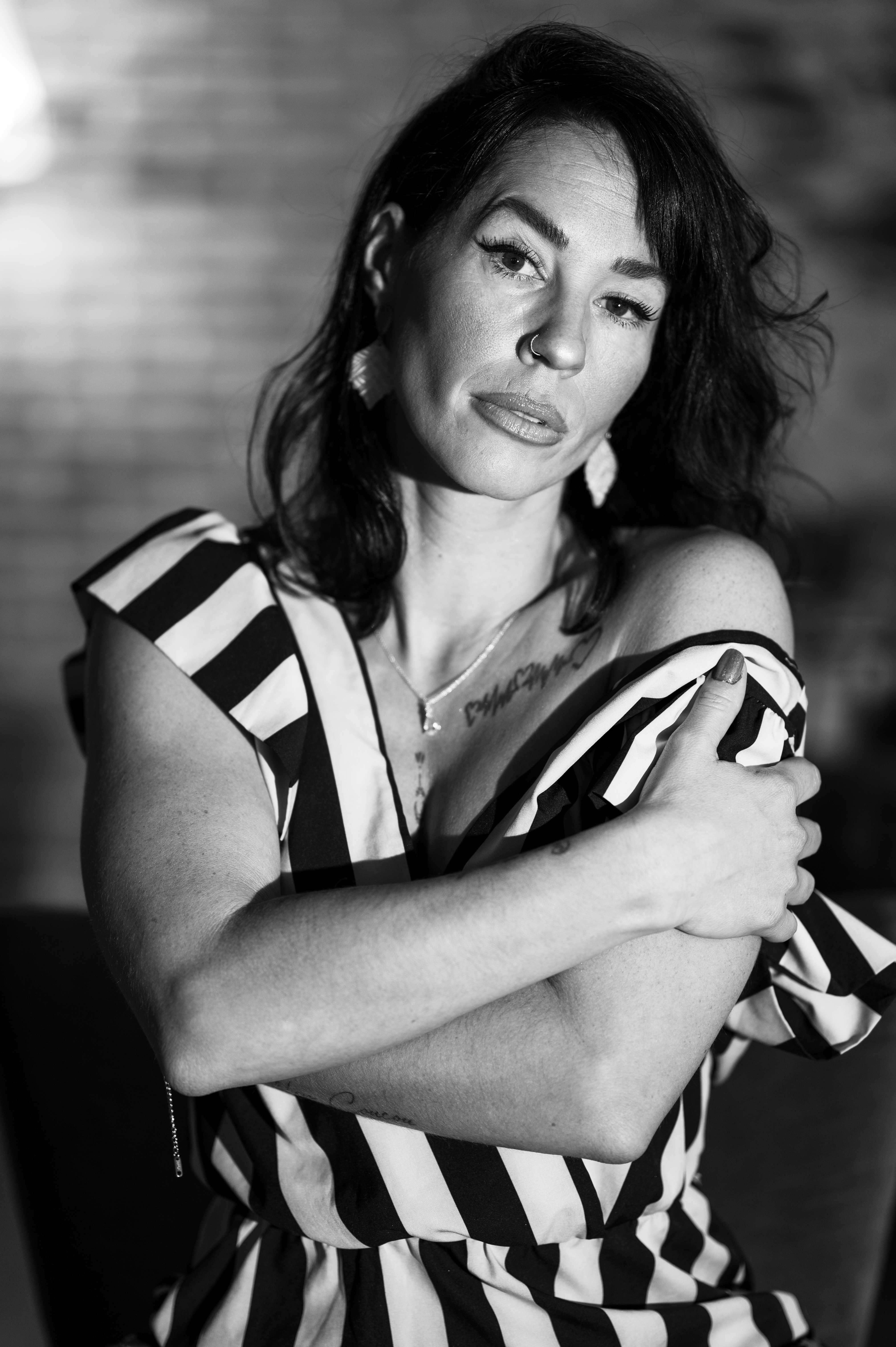 Femme en robe à rayures noires et blanches, photographiée en noir et blanc, les bras croisés sur la poitrine, portant un piercing au nez et des boucles d’oreilles pendantes, assise devant un mur de briques flou en arrière-plan.