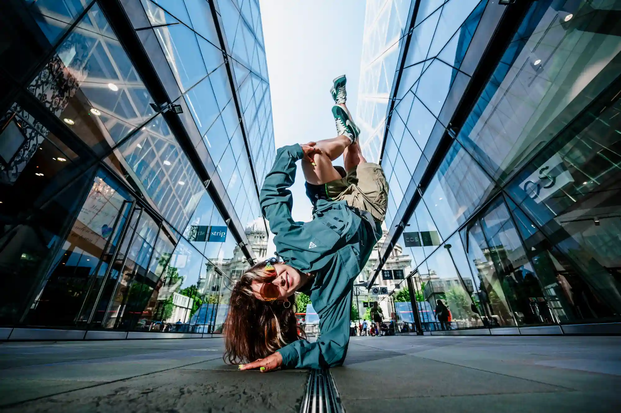 Une danseuse de breakdance réalise une figure impressionnante en équilibre sur un bras, dans une rue urbaine encadrée par deux immeubles en verre. Elle porte des lunettes de soleil, une veste bleu-vert, un short et des baskets. Elle sourit tout en maintenant sa pose dynamique.