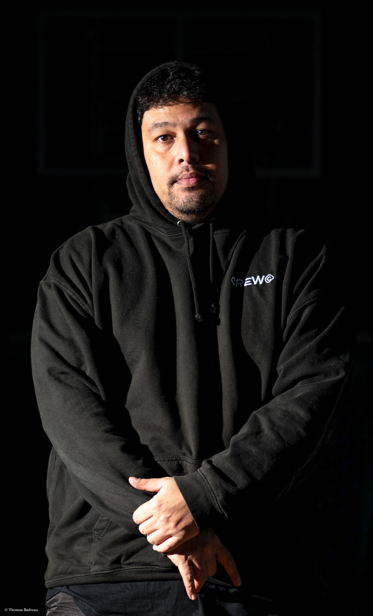 Homme portant un sweat à capuche noir avec l'inscription 'CREW@', debout dans l'obscurité avec une lumière dirigée sur son visage et le haut de son corps, posant avec les mains croisées devant lui.