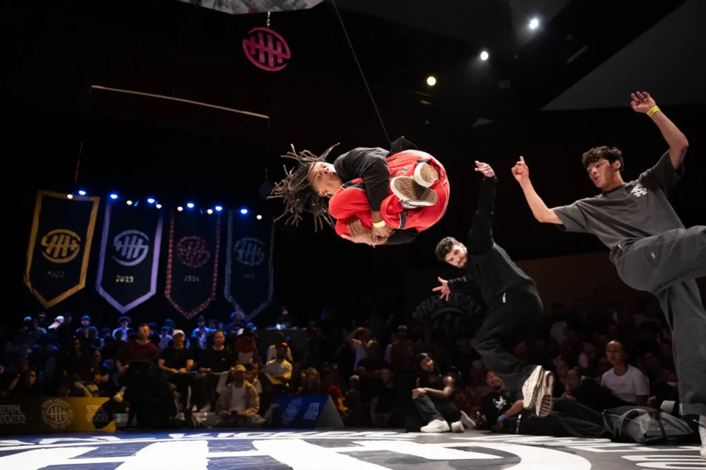 Lors d’un battle de breakdance en intérieur, un danseur aux dreadlocks effectue un saut acrobatique en position repliée, vêtu d’un pantalon rouge et d’un haut noir. Deux autres danseurs, habillés en noir et gris, l’encouragent avec enthousiasme. En arrière-plan, un public attentif et plusieurs bannières lumineuses décorent la scène.