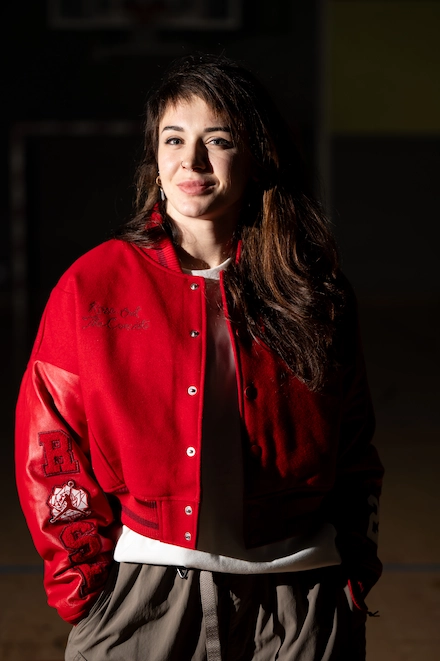 Jeune femme souriante portant une veste rouge de style universitaire, debout dans un gymnase faiblement éclairé