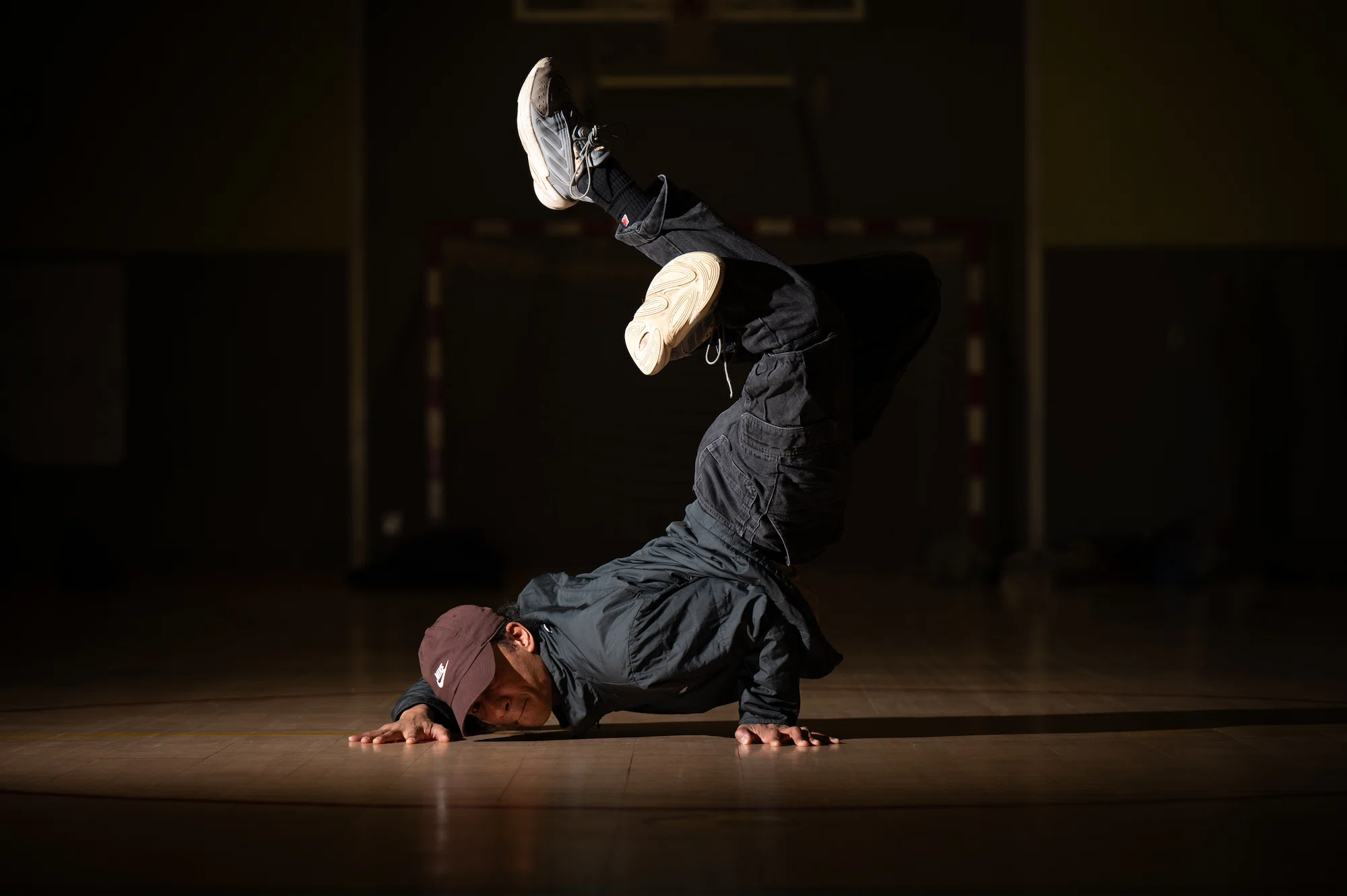 Un danseur de breakdance en casquette marron, veste foncée et pantalon cargo noir réalise un mouvement d’équilibre au sol en appui sur les mains, avec les jambes repliées et levées vers le haut. L’éclairage focalisé met en valeur le danseur dans une salle sombre.