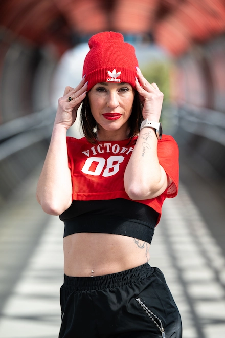 L’image montre une femme posant dans un tunnel piétonnier lumineux au design moderne. Elle porte un bonnet rouge Adidas, un crop top rouge avec l’inscription "VICTORY 08", un brassière noire en dessous, et un pantalon de sport noir à fermeture éclair. Elle touche son bonnet des deux mains tout en regardant directement l’objectif avec assurance. Elle a les lèvres rouges, un piercing au nombril, un tatouage visible sur le côté de la taille ainsi qu’un autre sur l’avant-bras droit. Elle porte également une Apple Watch. La scène dégage une énergie urbaine, sportive et confiante.