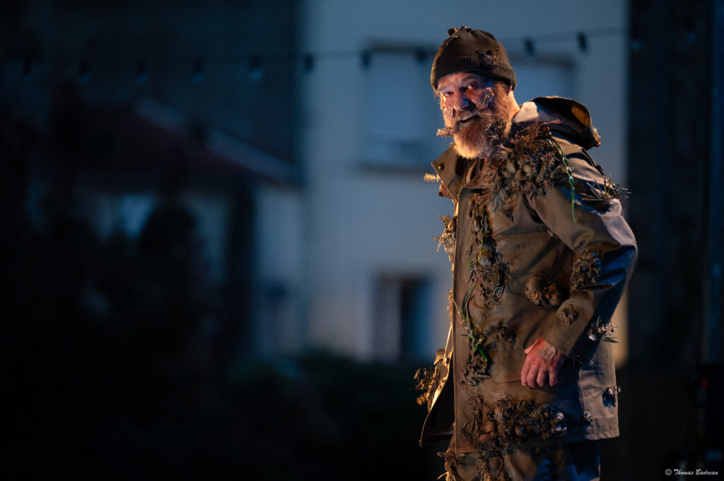 un homme se tient debout a droite de l'image déguisé avec un grand manteau remplis de champignons et d'herbe, il porte aussi un bonnet kaki et un micro bouche sur fond de décord école