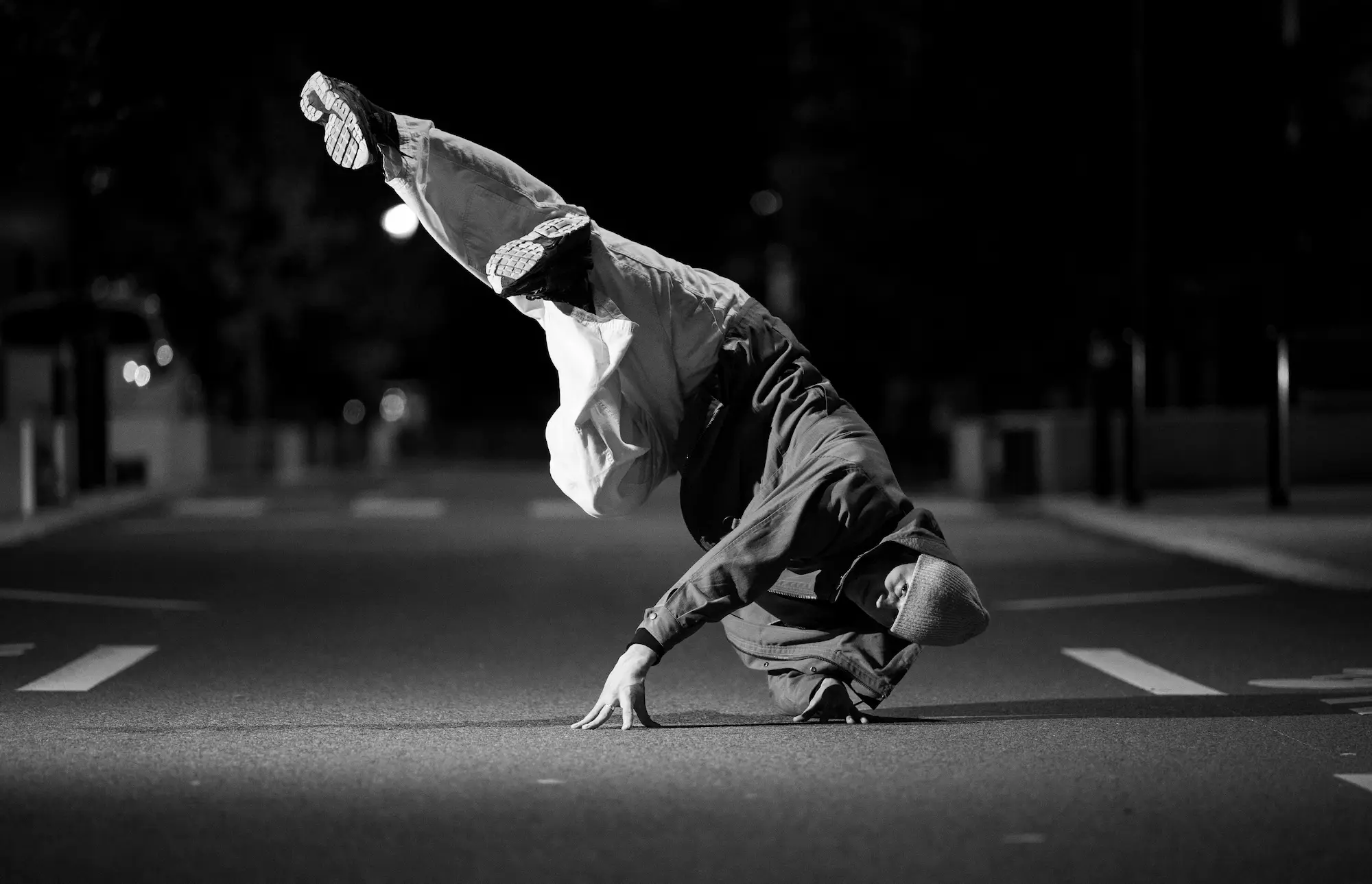 Un danseur de breakdance réalise une figure au sol en appui sur une main, dans une rue déserte la nuit, vêtu d'un bonnet, d'une veste et d'un pantalon ample.