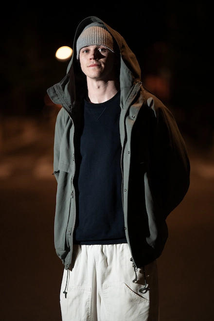 Un homme portant un bonnet gris, une veste verte à capuche et un pantalon clair pose de nuit dans une rue faiblement éclairée.