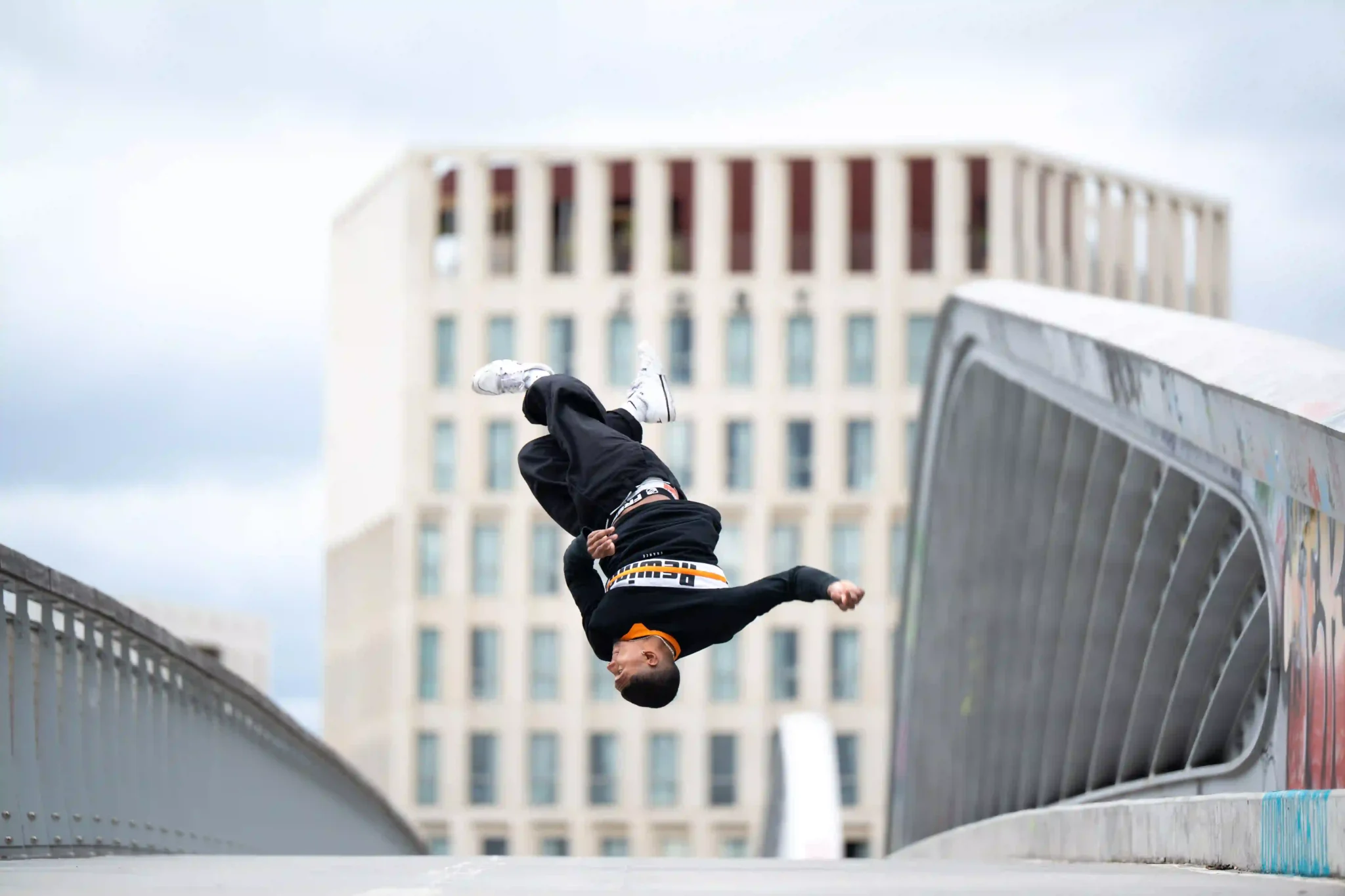 Un jeune homme en vêtements noirs, chaussures blanches, effectue un saut périlleux en l’air sur un pont urbain, avec un bâtiment moderne flou en arrière-plan.
