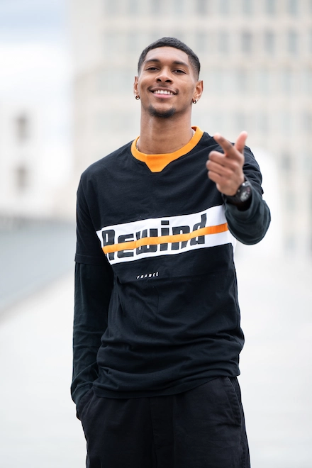 Un homme souriant, habillé d’un t-shirt noir avec l'inscription "Rewind" barrée d’une bande orange, pose en extérieur avec un geste de la main vers l’objectif.