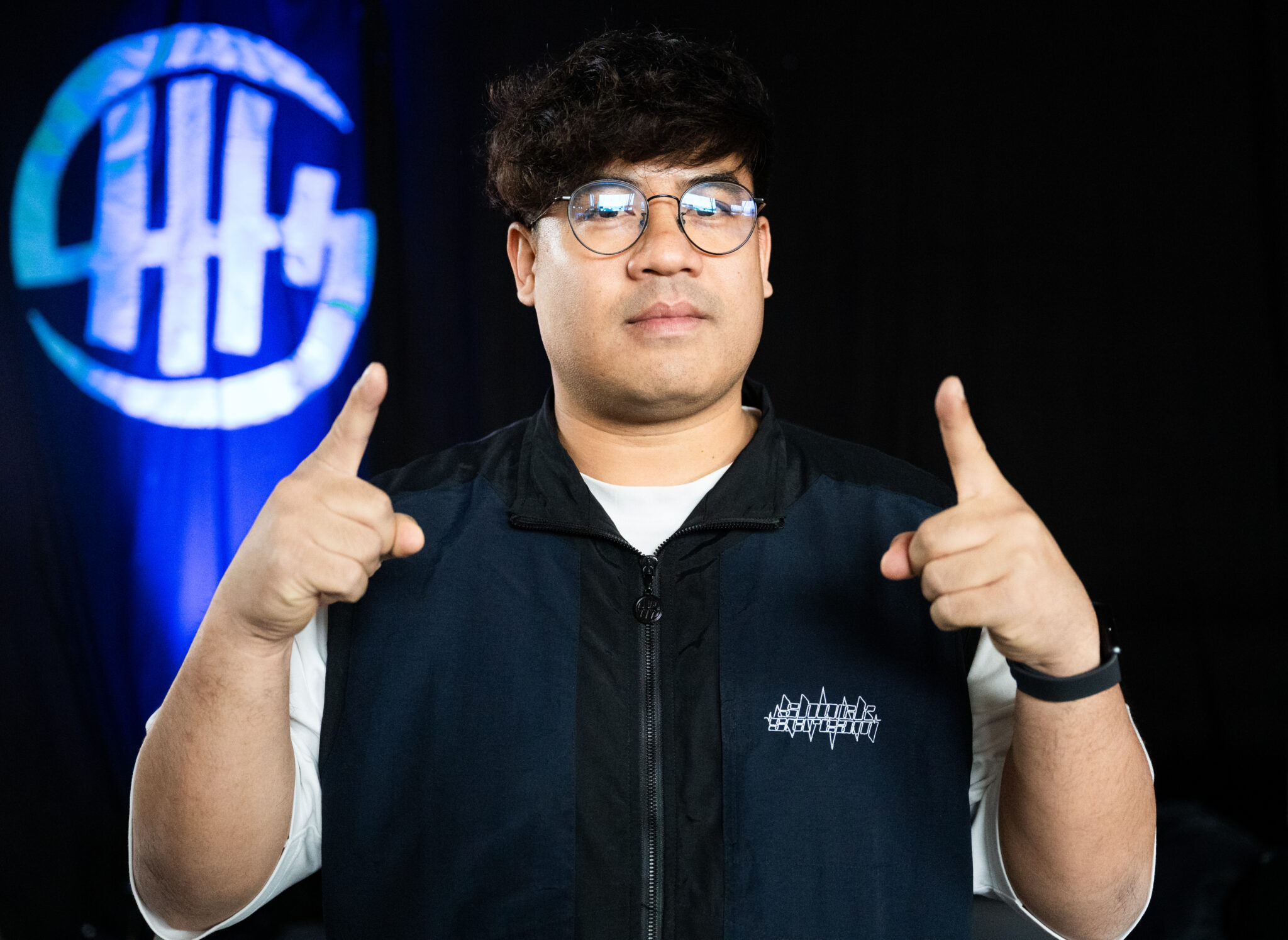 Homme portant des lunettes rondes, un t-shirt blanc et une veste noire avec un logo graphique, pointant vers le haut avec les deux index, debout devant un fond sombre avec un logo lumineux bleu et blanc en arrière-plan