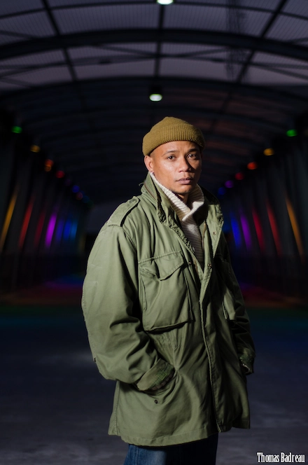 Homme portant une veste militaire verte, un bonnet marron et un pull clair, posant dans un tunnel illuminé par des lumières colorées, avec une expression sérieuse et les mains dans les poches.