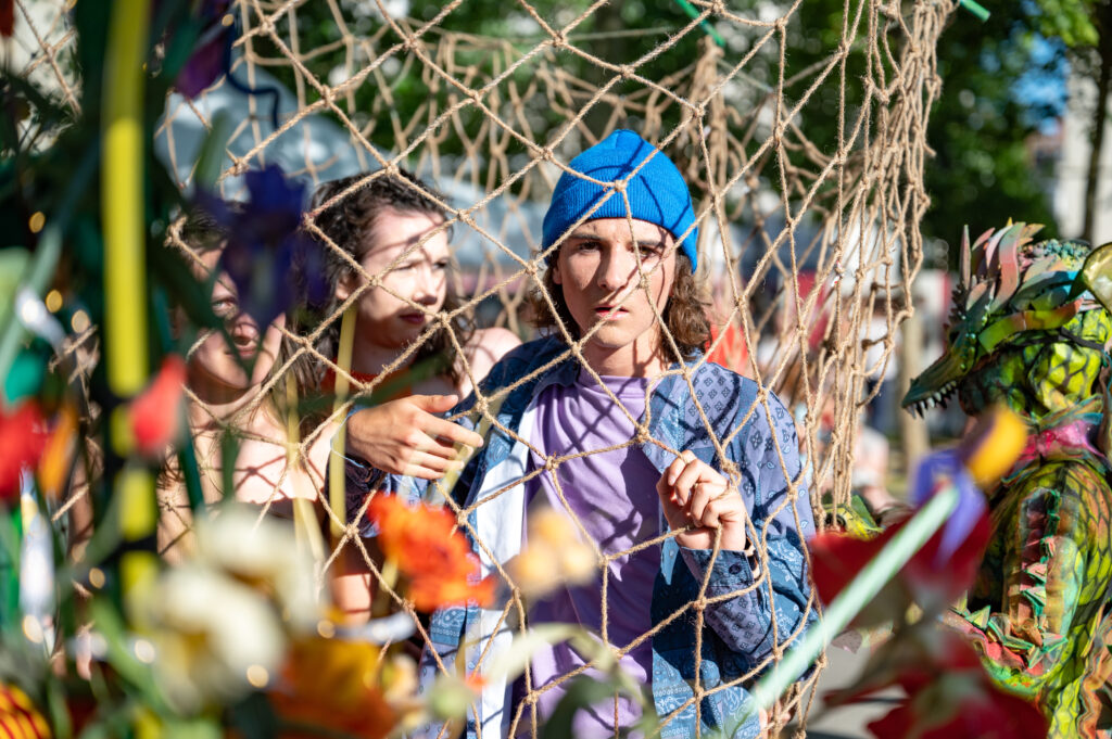 Un jeune homme portant un bonnet bleu et une chemise bleue à motifs est vu à travers un filet de corde. Il regarde directement l’objectif avec une expression sérieuse. Derrière lui, une femme au visage partiellement visible et une personne habillée en costume coloré ressemblant à un dragon ou une créature fantastique apparaissent floues. Au premier plan, des éléments colorés et flous encadrent la scène, ajoutant de la profondeur à la photo.