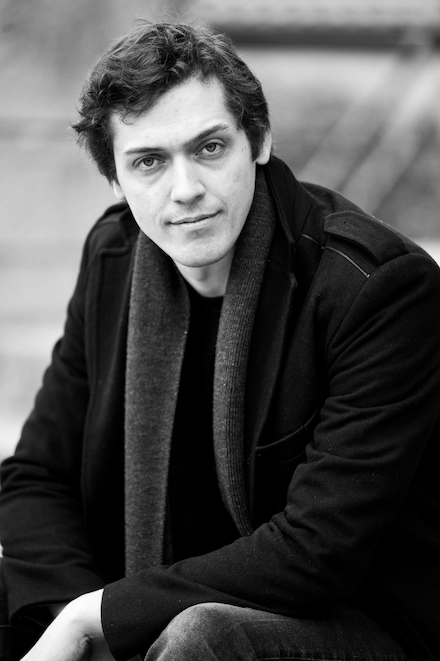 Portrait en noir et blanc d’un homme aux cheveux bouclés, portant un manteau et une écharpe, regardant directement l’objectif avec une expression calme.