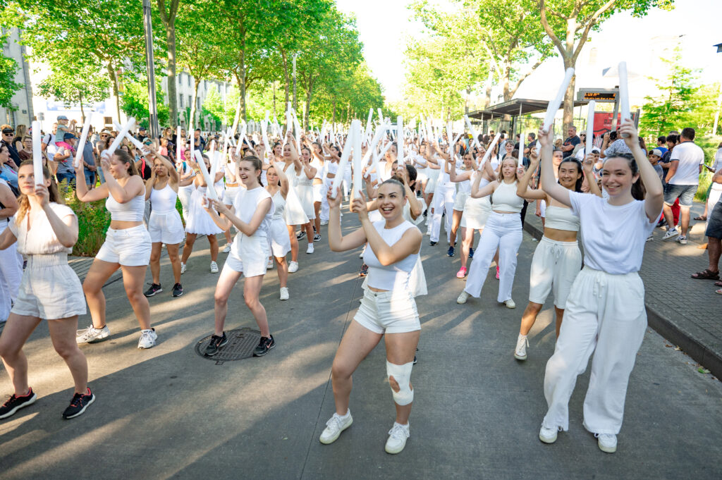 Une grande foule de jeunes femmes habillées en blanc participent à une performance en plein air. Elles tiennent toutes des bâtons blancs qu’elles lèvent en l'air en même temps, semblant danser ou suivre une chorégraphie. Elles sont sur une route bordée d’arbres verts, avec des spectateurs qui les regardent sur les côtés. L’ambiance est joyeuse et ensoleillée.