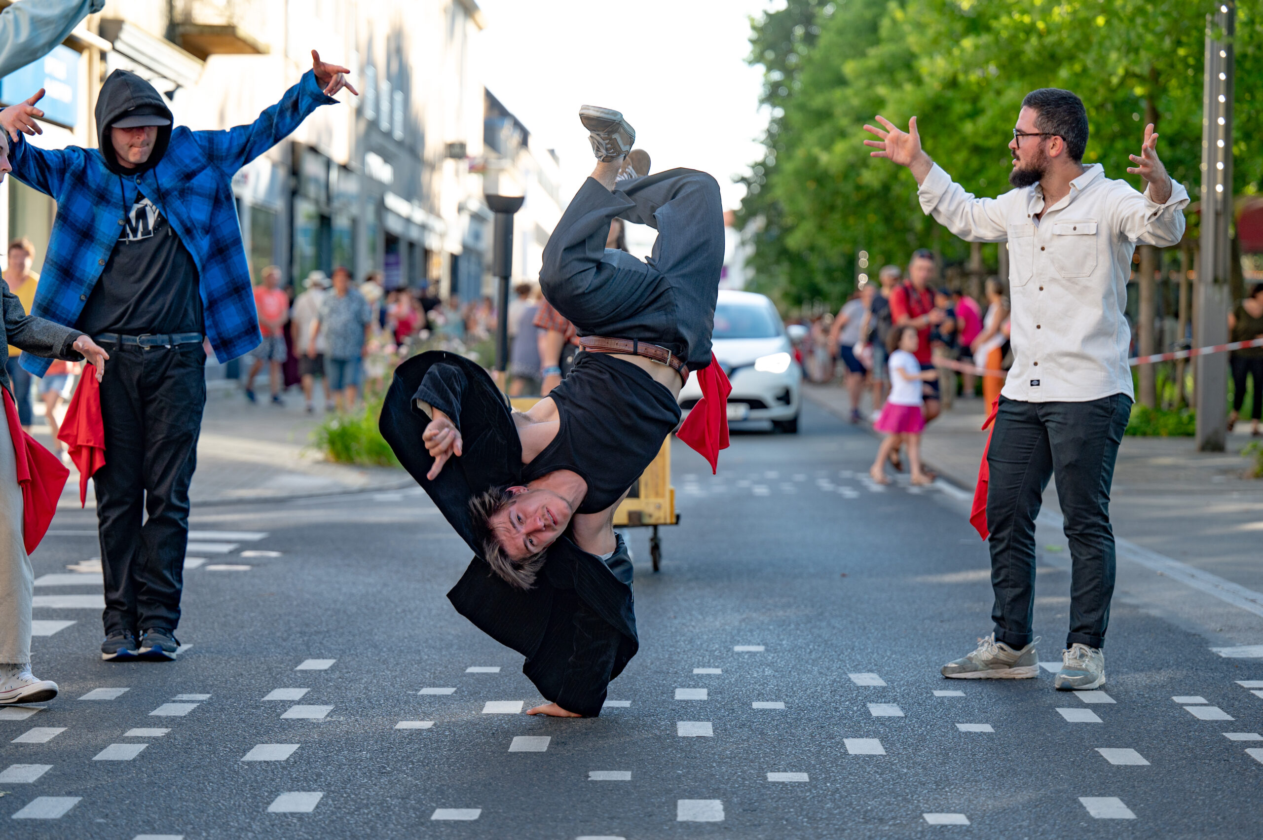 Un groupe de danseurs de rue en pleine performance sur une route urbaine. Un danseur au centre est en équilibre sur une main, réalisant un mouvement de breakdance impressionnant. Il porte un pantalon gris, un débardeur noir et une veste noire ouverte. Autour de lui, d’autres danseurs encouragent et posent des gestes dynamiques. L’un porte une chemise blanche, un autre une veste à carreaux bleue avec un sweat à capuche noire. En arrière-plan, on aperçoit des passants, des arbres et des bâtiments.