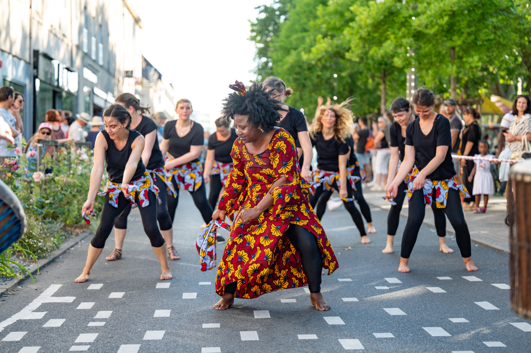 Une troupe de danseurs en plein spectacle en extérieur sur une rue piétonne. Au centre, une femme portant une robe colorée rouge avec des motifs jaunes et noirs danse de manière expressive, tenant un tissu assorti. Autour d'elle, plusieurs danseuses vêtues de leggings noirs et de t-shirts noirs, avec des tissus colorés noués à la taille, exécutent des mouvements synchronisés. Des spectateurs sont visibles en arrière-plan, ainsi que des arbres et des bâtiments.