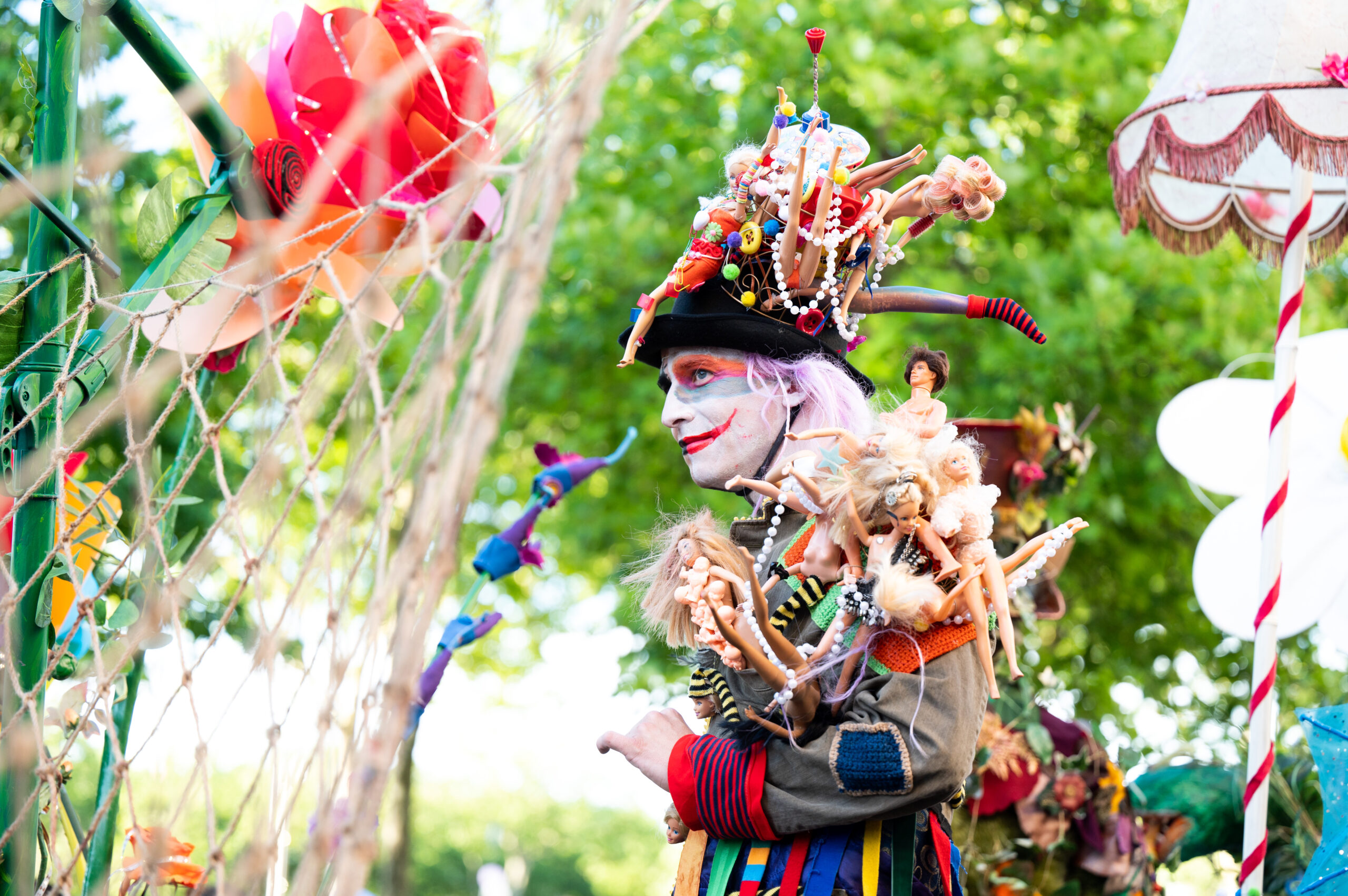 Un artiste déguisé et maquillé déambule dans un décor festif et coloré en plein air. Il porte un costume extravagant orné de poupées, perles, peluches et objets variés, avec un chapeau spectaculaire chargé de figurines et de bibelots. Son visage est maquillé de façon théâtrale, avec des couleurs vives et un sourire exagéré rappelant un clown ou un personnage de carnaval. L’arrière-plan montre des arbres verts, des décorations fantaisistes et des lampes rétro, suggérant une ambiance de festival artistique ou de parade créative.