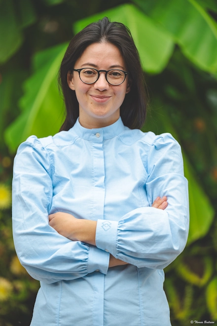 Femme souriante portant des lunettes et une chemise bleu clair, les bras croisés, posant devant un fond de feuilles vertes.