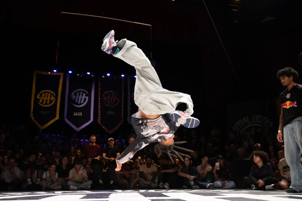 Un danseur de breakdance est en plein saut acrobatique, tournant la tête en bas avec les jambes fléchies dans les airs. Il porte un pantalon clair, un sweat à manches longues imprimé et des baskets colorées. En arrière-plan, un public attentif observe la performance dans une salle sombre avec des bannières suspendues indiquant les années 2022, 2023, 2024.