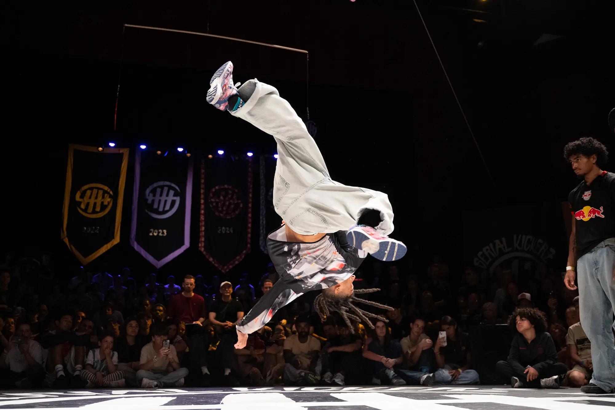 Un danseur de breakdance est en plein saut acrobatique, tournant la tête en bas avec les jambes fléchies dans les airs. Il porte un pantalon clair, un sweat à manches longues imprimé et des baskets colorées. En arrière-plan, un public attentif observe la performance dans une salle sombre avec des bannières suspendues indiquant les années 2022, 2023, 2024.