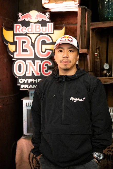 Un homme portant une casquette Red Bull et un sweat noir "Reiginal" pose devant une enseigne lumineuse du Red Bull BC One à Orphéon, France, un événement international de danse hip-hop.