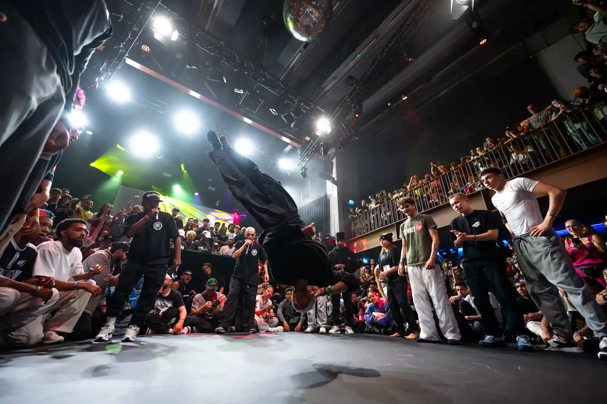 Un danseur de breakdance est en plein mouvement acrobatique, exécutant un saut ou une figure sur la tête, au centre d’un cercle formé par un public nombreux et enthousiaste. La scène est illuminée par des projecteurs puissants, avec des effets de lumière colorée en arrière-plan. L’événement semble se dérouler dans une salle de concert ou une boîte de nuit, avec un étage supérieur bondé de spectateurs regardant la scène. L’ambiance est vibrante, dynamique et électrique, typique d’une compétition ou d’un battle de danse urbaine.