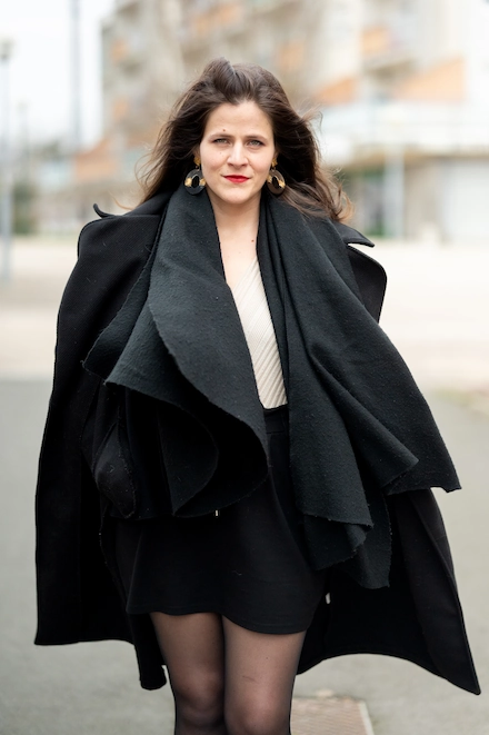 Femme brune marchant à l’extérieur, portant un manteau noir ample, une jupe courte noire, un haut clair plissé et des collants noirs. Elle a les cheveux lâchés, porte des boucles d’oreilles rondes dorées et affiche une expression confiante.