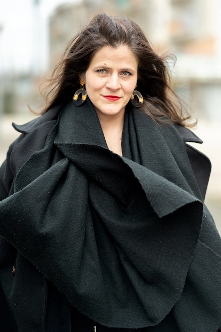 Femme brune marchant à l’extérieur, portant un manteau noir ample, une jupe courte noire, un haut clair plissé et des collants noirs. Elle a les cheveux lâchés, porte des boucles d’oreilles rondes dorées et affiche une expression confiante.