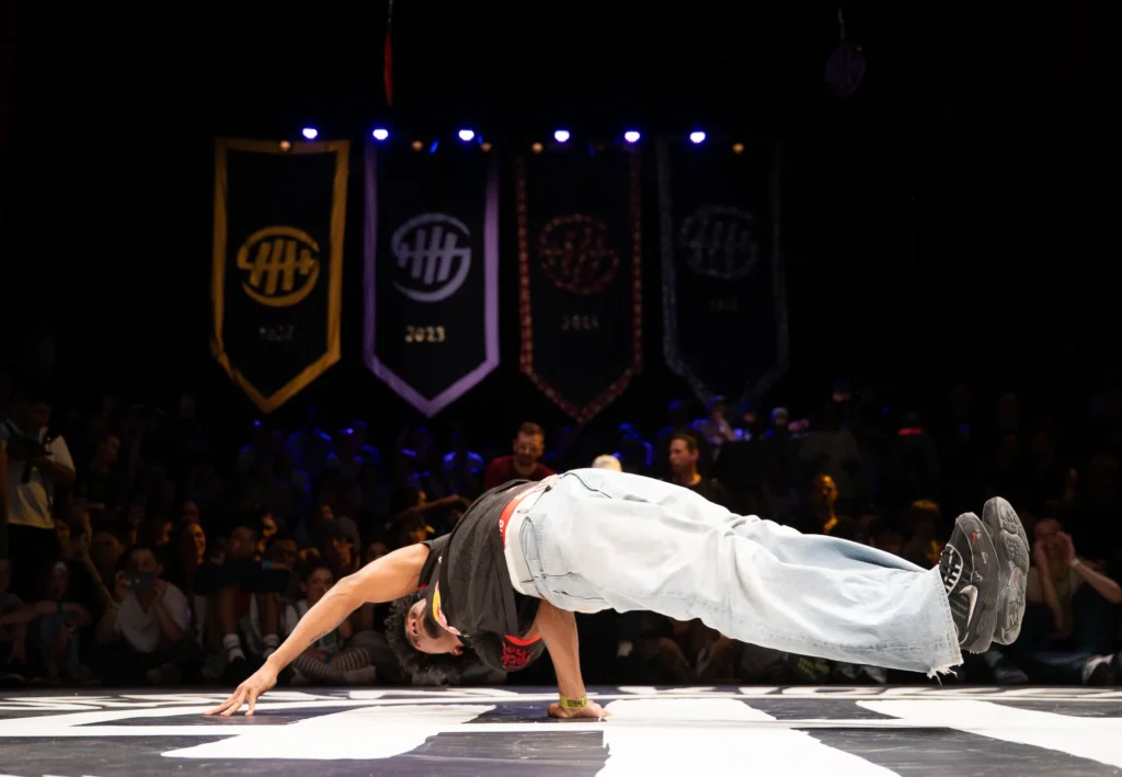 Un danseur de breakdance réalise un mouvement de force en appui sur une main, son corps parallèle au sol et ses jambes tendues. Il porte un débardeur noir et un jean clair. En arrière-plan, un public nombreux observe la performance dans une salle sombre, avec plusieurs bannières suspendues affichant des logos et des années.