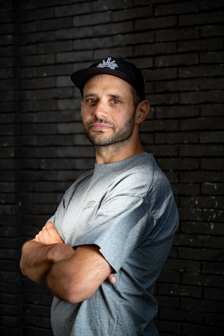 Homme debout devant un mur de briques sombres, les bras croisés. Il porte une casquette noire et un t-shirt gris, et regarde l’objectif avec un air calme et assuré. La lumière met en valeur son visage et le haut de son corps.