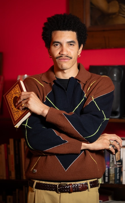 Homme debout dans une bibliothèque, tenant un livre à la couverture ornée et un verre à la main, vêtu d’un pull à losanges marron et bleu marine, avec une ceinture tressée et des bijoux.