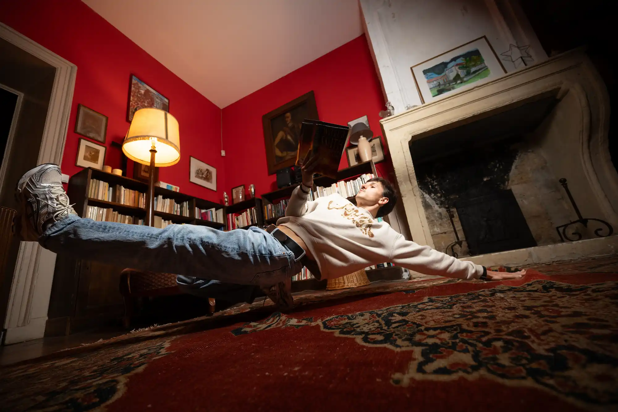 Un jeune homme en sweat-shirt blanc et jean réalise un équilibre latéral impressionnant dans un salon rouge, tenant un livre dans une main, avec une bibliothèque, une lampe et une cheminée en arrière-plan.
