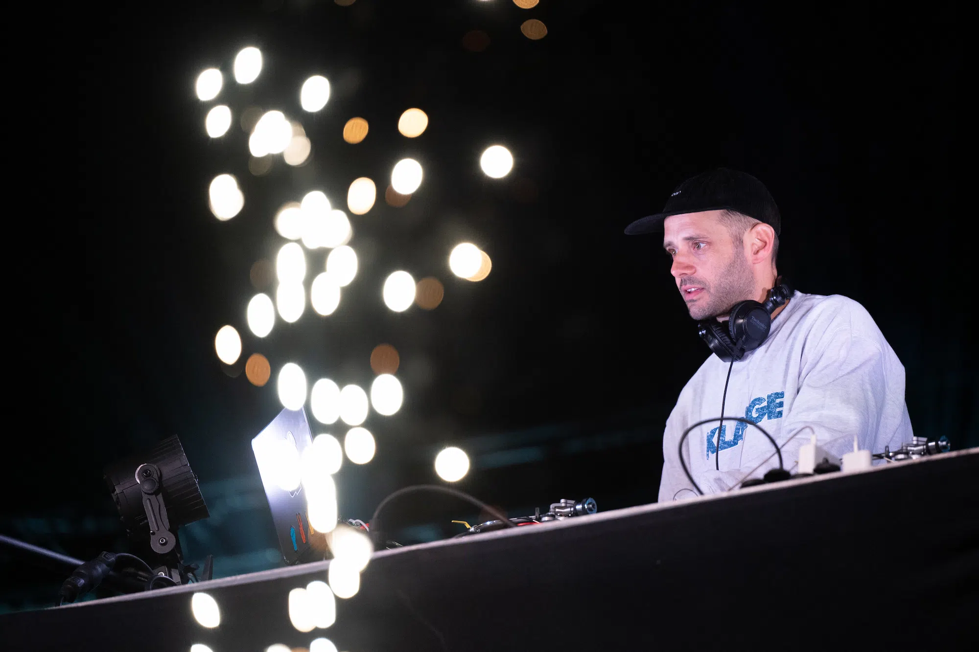 Un DJ concentré travaille derrière sa console lors d’un événement nocturne. Il porte un sweat gris avec un casque audio autour du cou et une casquette noire. Devant lui, des lumières floues et brillantes créent une ambiance festive.