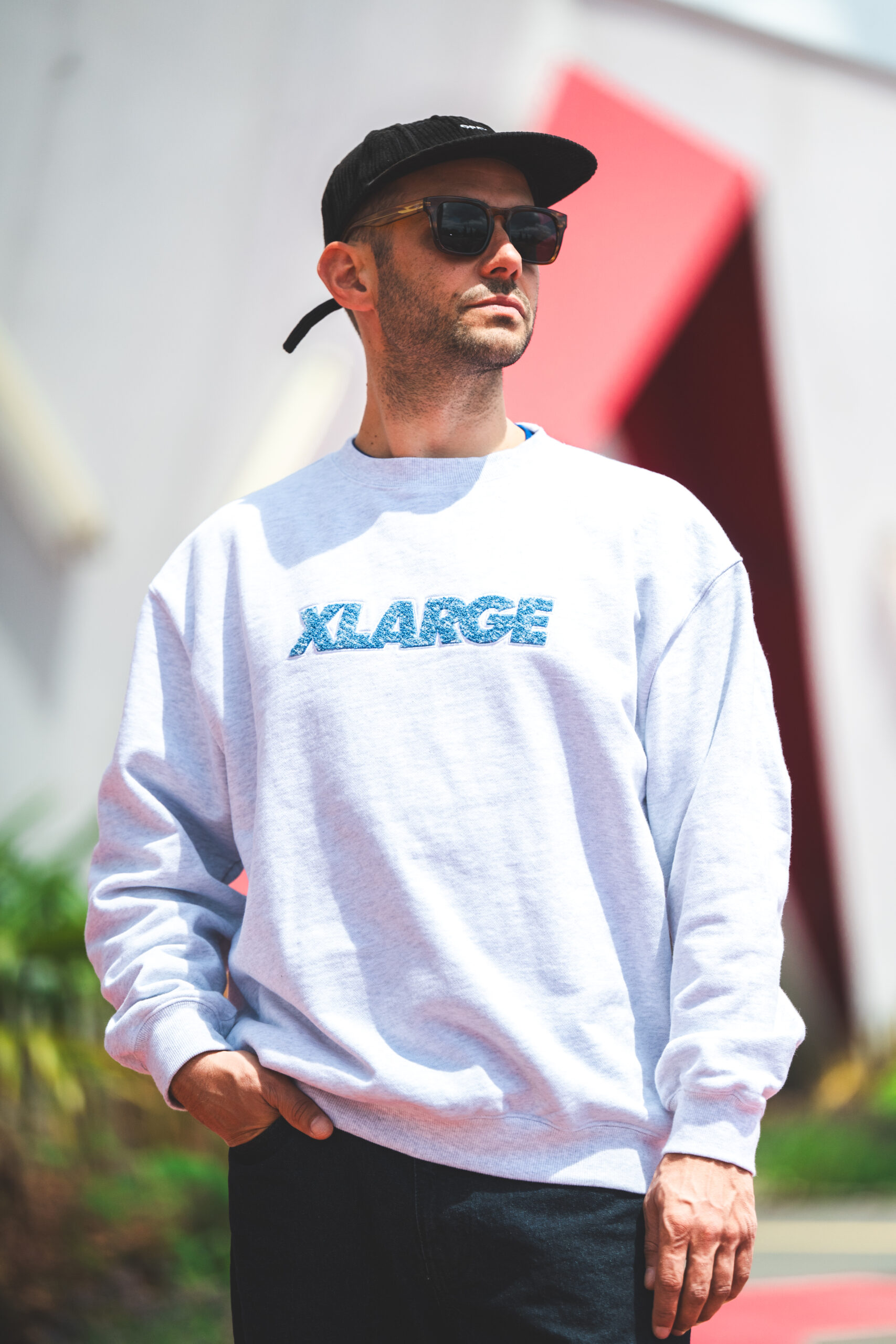 Homme portant un sweat gris avec l'inscription 'XLARGE' en bleu pailleté, une casquette noire à l'envers et des lunettes de soleil, posant à l'extérieur devant un bâtiment moderne aux formes géométriques rouges et blanches.