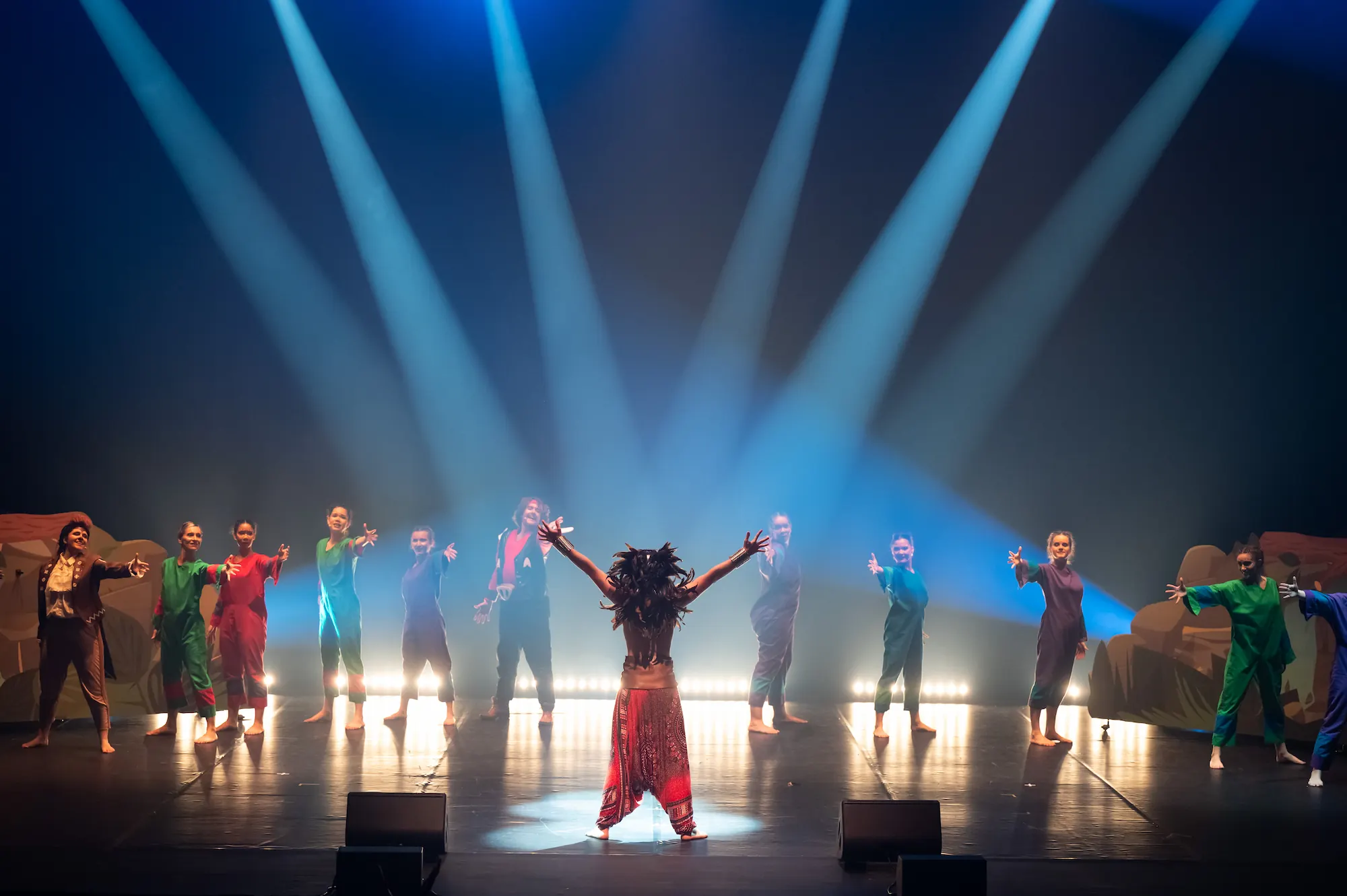 Groupe de danseurs et danseuses sur scène sous des projecteurs bleus, formant un demi-cercle autour d'une personne au centre, de dos, les bras levés. La scène est éclairée de manière théâtrale avec une atmosphère dramatique. Tous sont pieds nus et portent des costumes colorés.