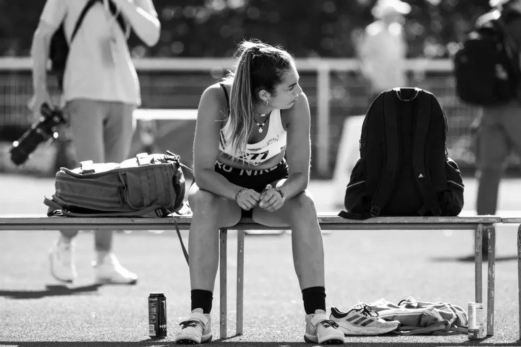 photo en noir et blanc d'une athlète assis sur un banc avec ses sacs posé a sur le banc. Au second plan gauche, un photographe