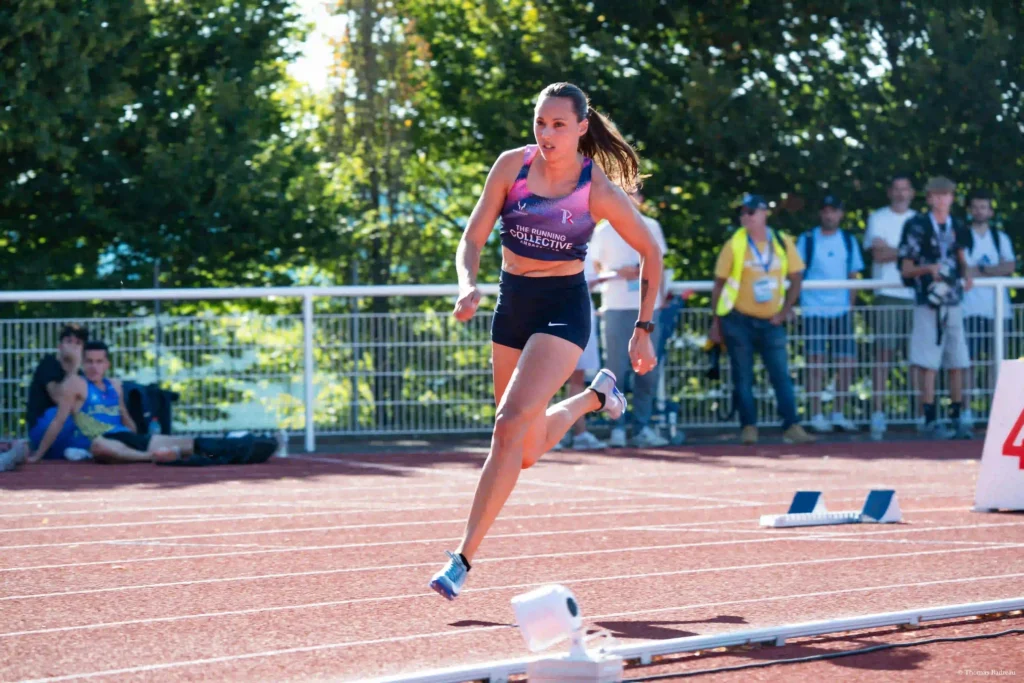 athlète femme courant sur la piste d'athlétisme en plein virage avec une tenue rose et noir sur fond de public et d'arbre