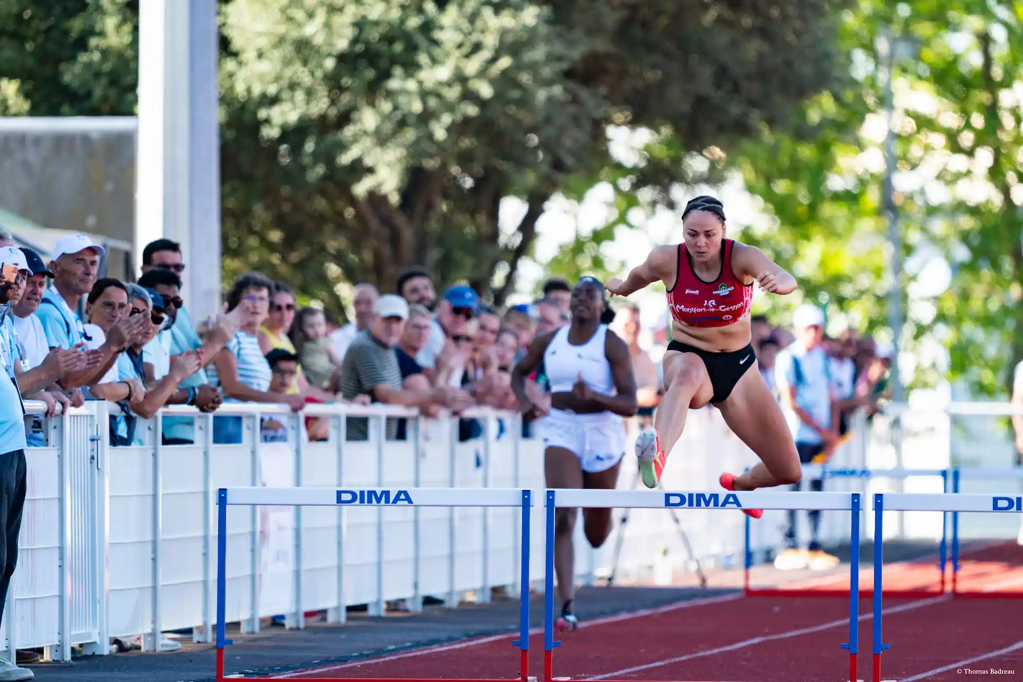 Une athlète en plein saut au-dessus d’une haie lors d’une course d’athlétisme, sous les applaudissements d’un public nombreux derrière une barrière. Elle porte une tenue rouge et noire, concentrée sur son effort, tandis qu’une autre concurrente en blanc suit derrière elle.