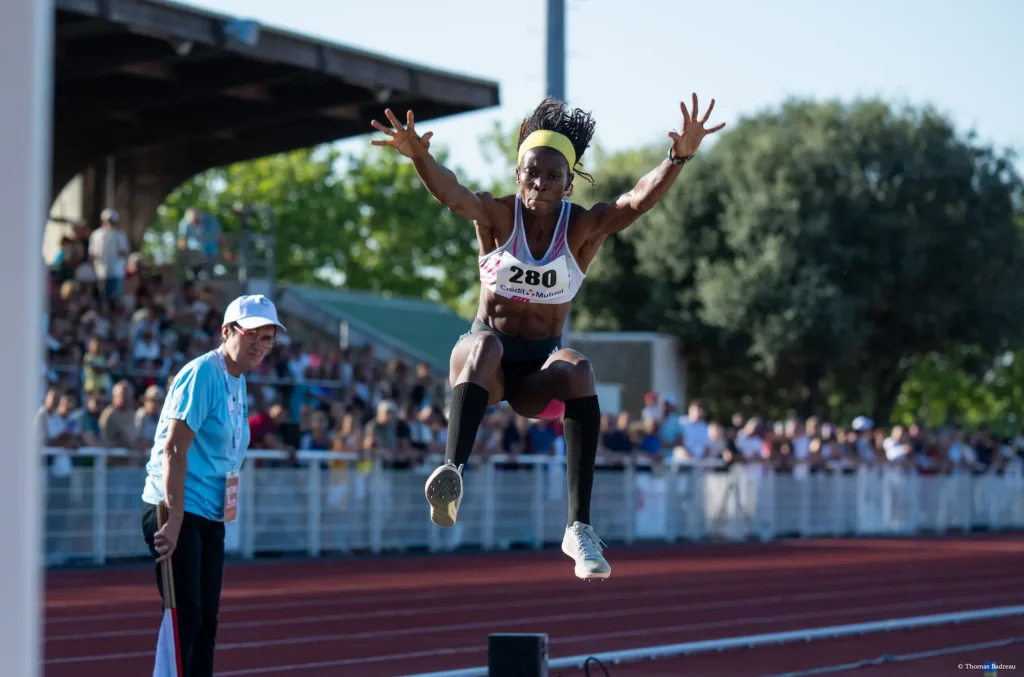 Athlète en plein saut en longeur sur une piste d'athlètisme avec à sa droite un juge et en fond un public dans un stade