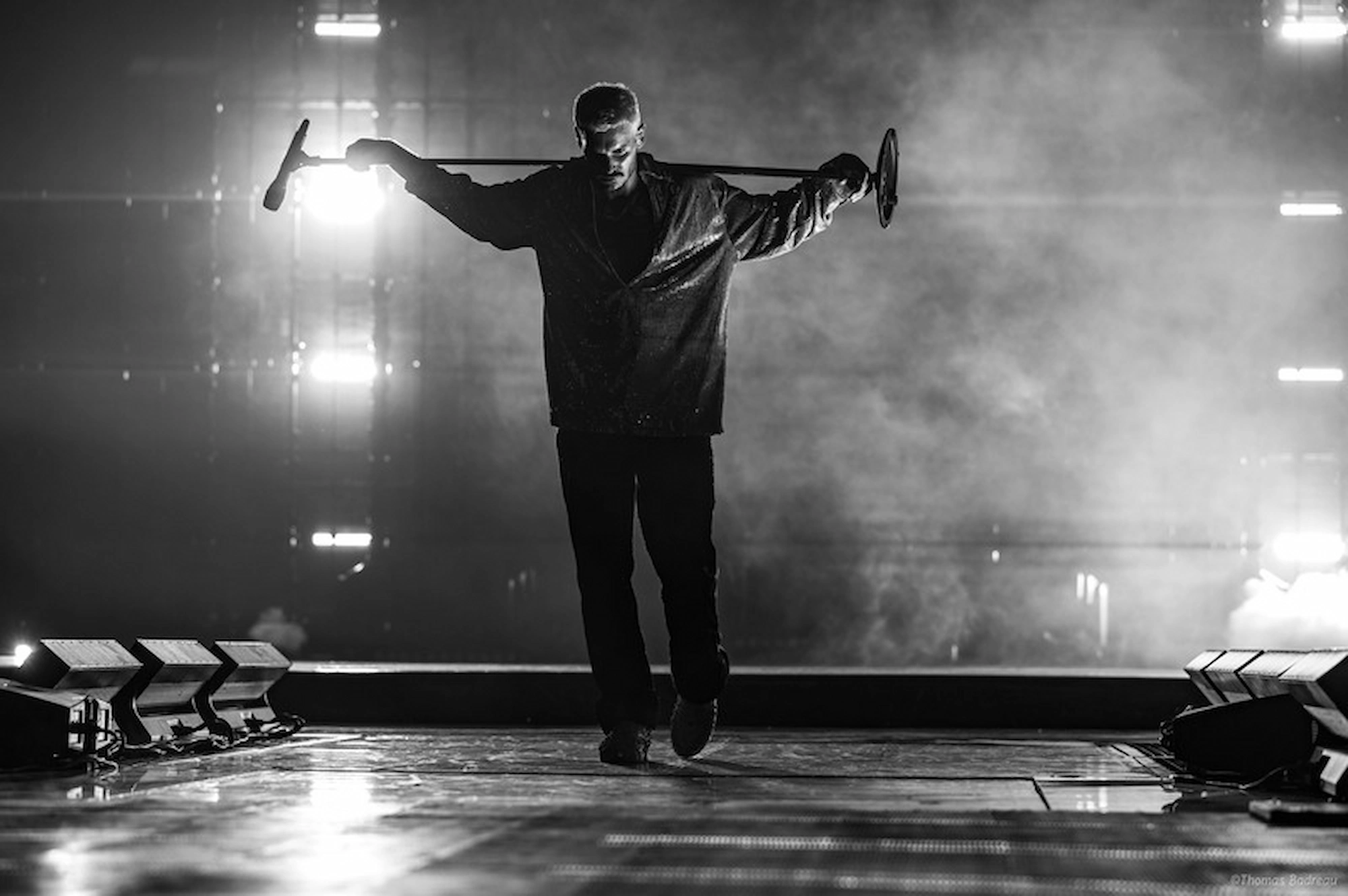 L’image en noir et blanc montre Matt Pokora sur scène, capturé en pleine performance dramatique. Il marche en tenant un long micro ou une barre sur ses épaules, les bras étendus de chaque côté. L’arrière-plan est plongé dans une atmosphère brumeuse, illuminée par des lumières vives et des projecteurs qui créent une silhouette marquante et théâtrale. Le sol brillant reflète la lumière, ajoutant de la profondeur à la scène. L’ensemble dégage une ambiance intense et cinématographique, évoquant une forte présence scénique et émotionnelle.