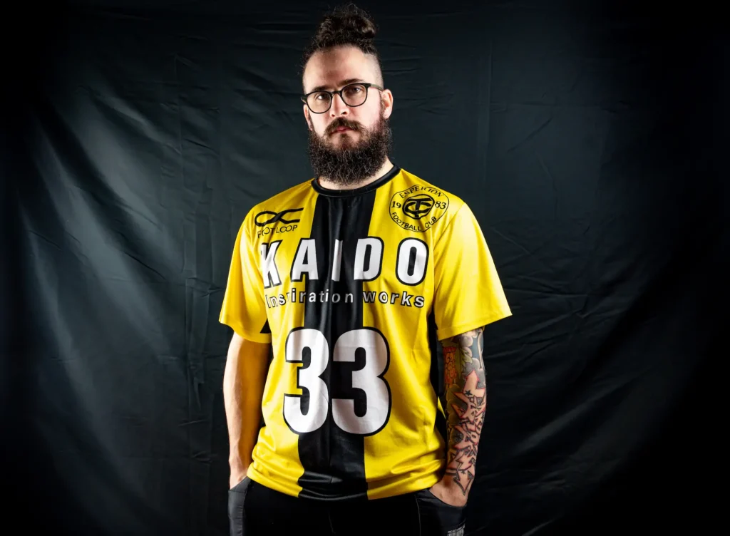 L'image montre Thomas Badreau debout devant un fond noir. Il porte un t-shirt de style sportif jaune et noir avec les inscriptions "KAIDO inspiration works" et le chiffre "33" en grand. Sur le haut du t-shirt, on peut lire également "FOOT LOOP" et "ESPERI TOY FOOTBALL CLUB 1983". L’homme a les cheveux attachés en chignon, porte des lunettes rondes et a des tatouages visibles sur son bras droit. Son expression est neutre, voire sérieuse. L’éclairage met bien en valeur les détails du visage, des tatouages et du t-shirt.